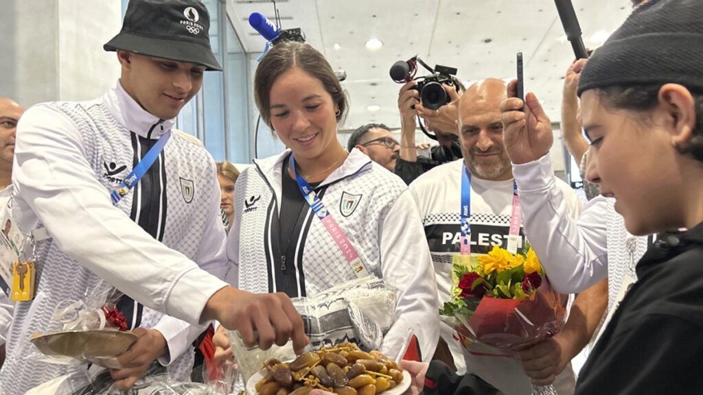 Los deportistas palestinos Yazan Al Bawwab y Valerie Tarazi toman dátiles ofrecidos por un niño a su llegada al aeropuerto de París, el jueves 25 de julio de 2024 (AP Foto/Megan Janetsky)